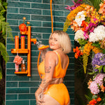 Person in orange swimsuit standing under an orange shower with a floral arrangement on a green tiled wall.