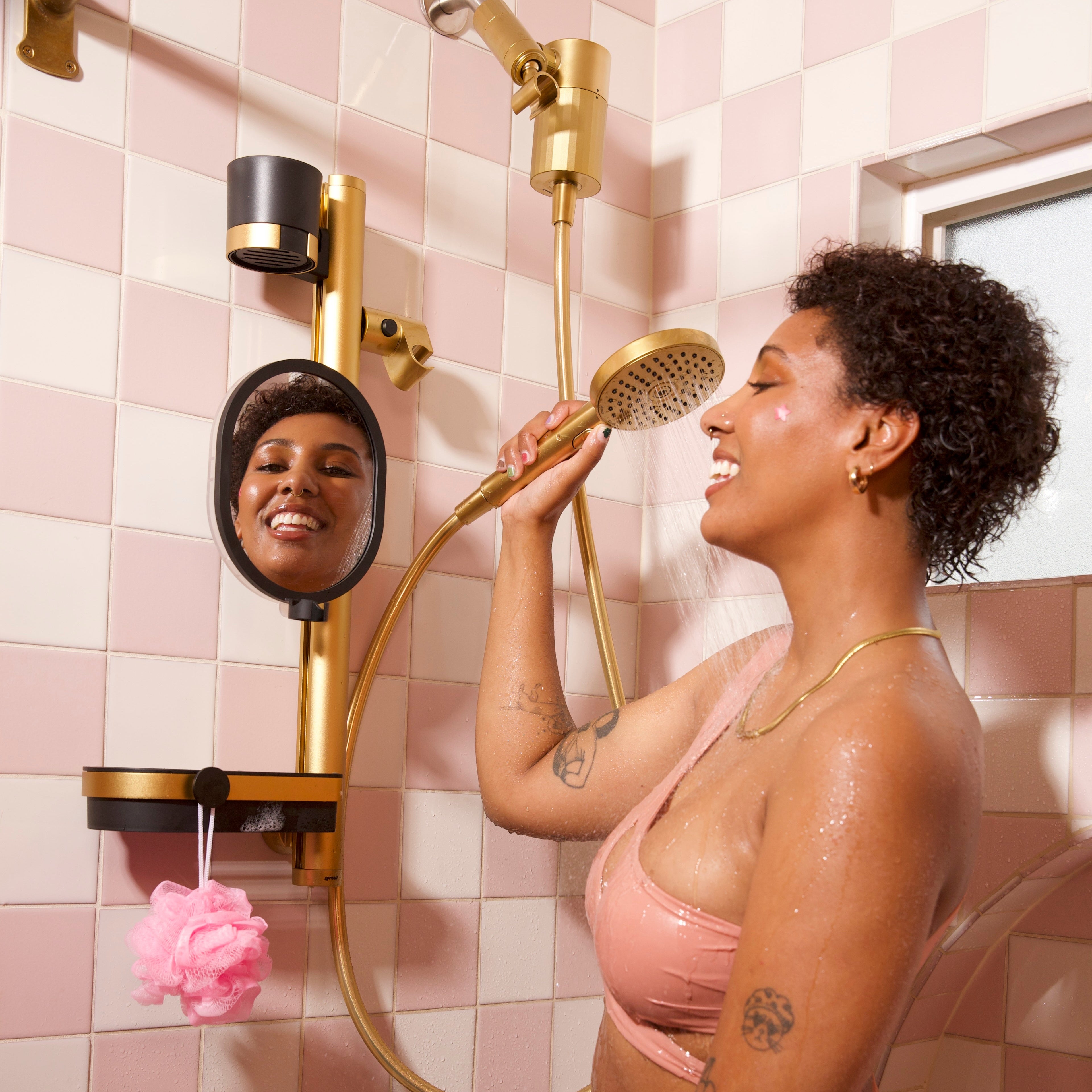 A woman in a pink bikini stands in a pink-and-gold tiled shower, holding a gold handheld shower head. A round wall mirror on the left reflects her face, and a pink loofah hangs from the shower fixture.