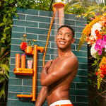 Man in orange swim shorts standing under a shower with a tropical setting