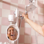 A person using a silver shower head with water running from a pink and white tiled wall bathroom setup, with a small round mirror and toiletry bottles visible.