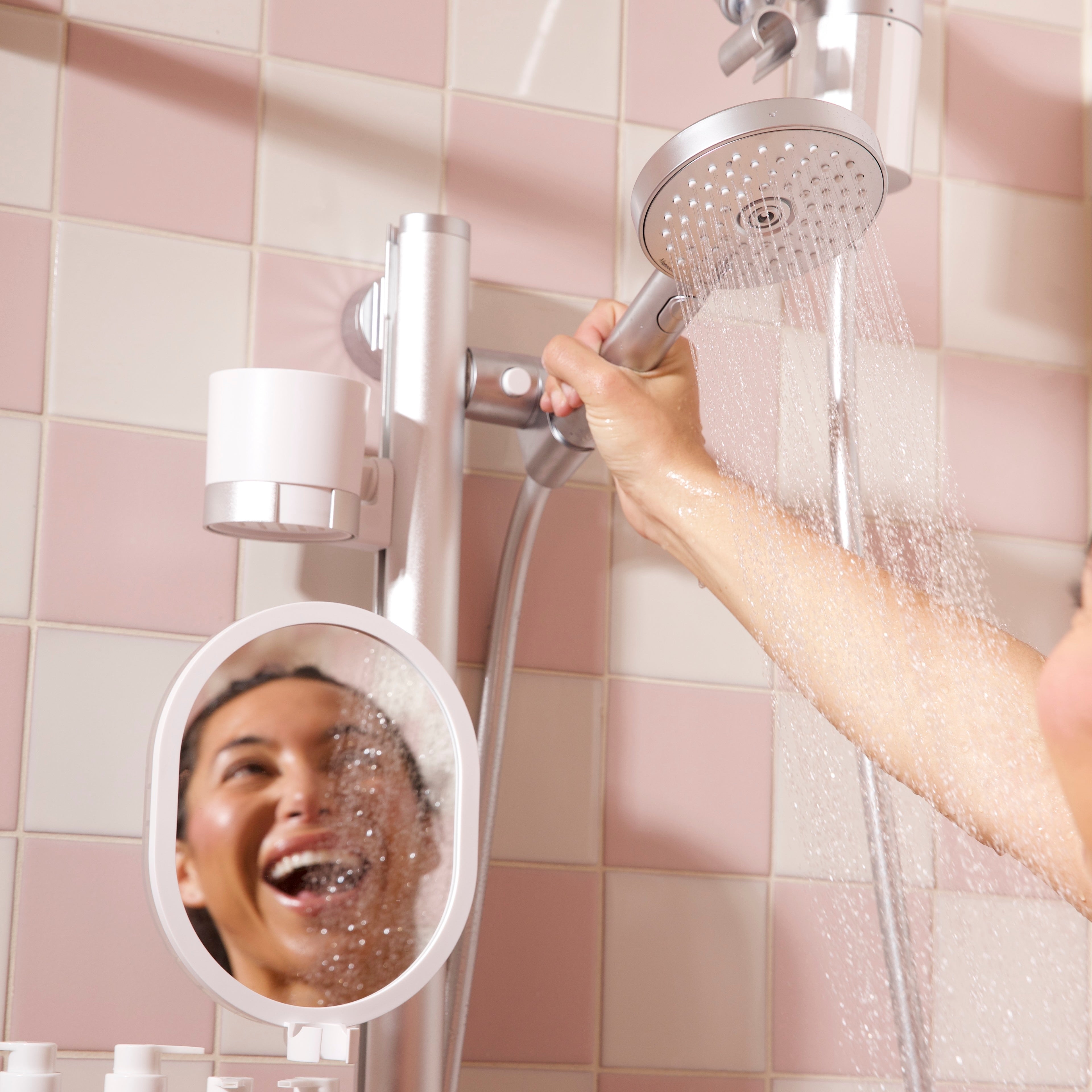 A person using a silver shower head with water running from a pink and white tiled wall bathroom setup, with a small round mirror and toiletry bottles visible.