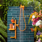 Outdoor shower with blue tiled wall, orange shower head and fixtures, surrounded by greenery and flowers.
