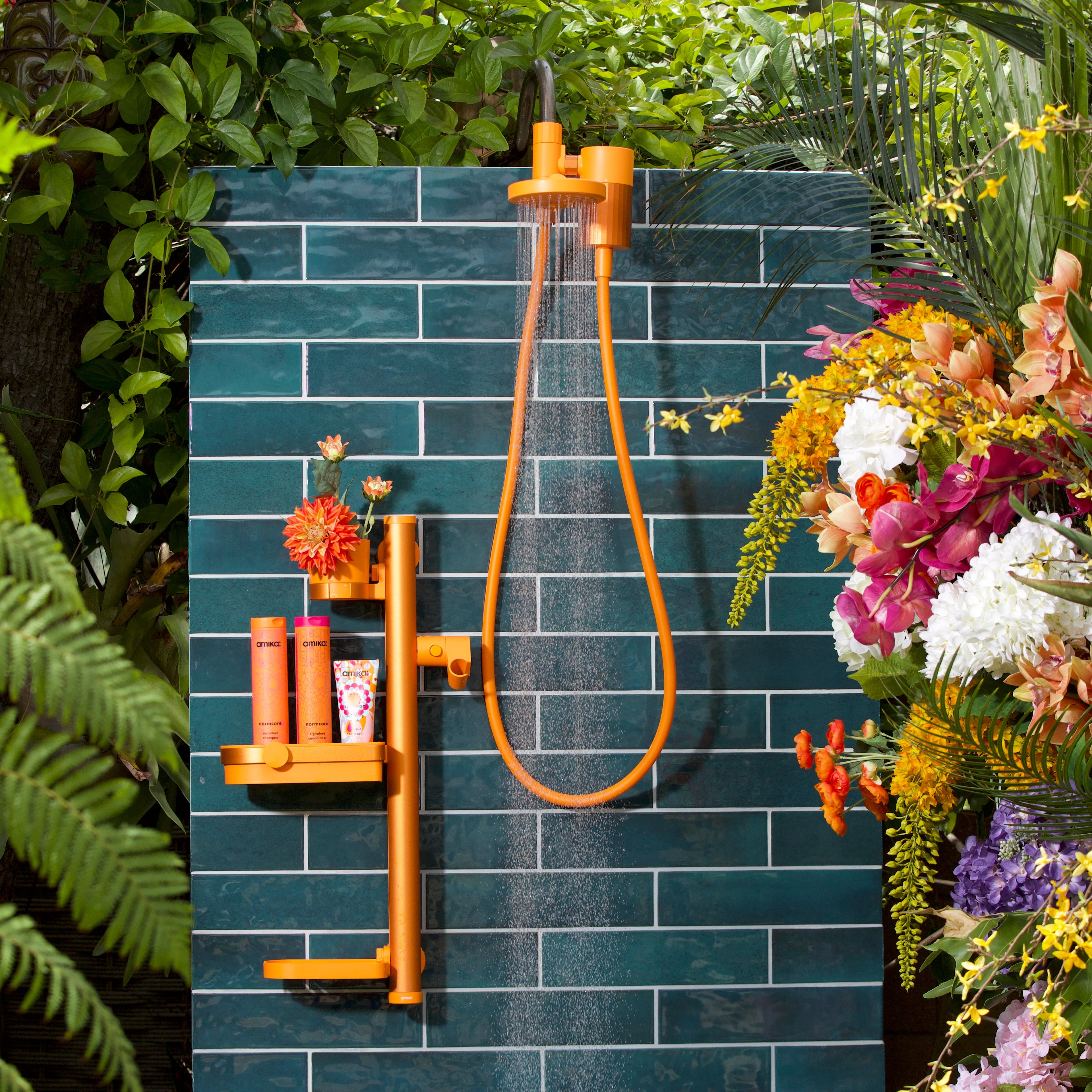 Outdoor shower with blue tiled wall, orange shower head and fixtures, surrounded by greenery and flowers.