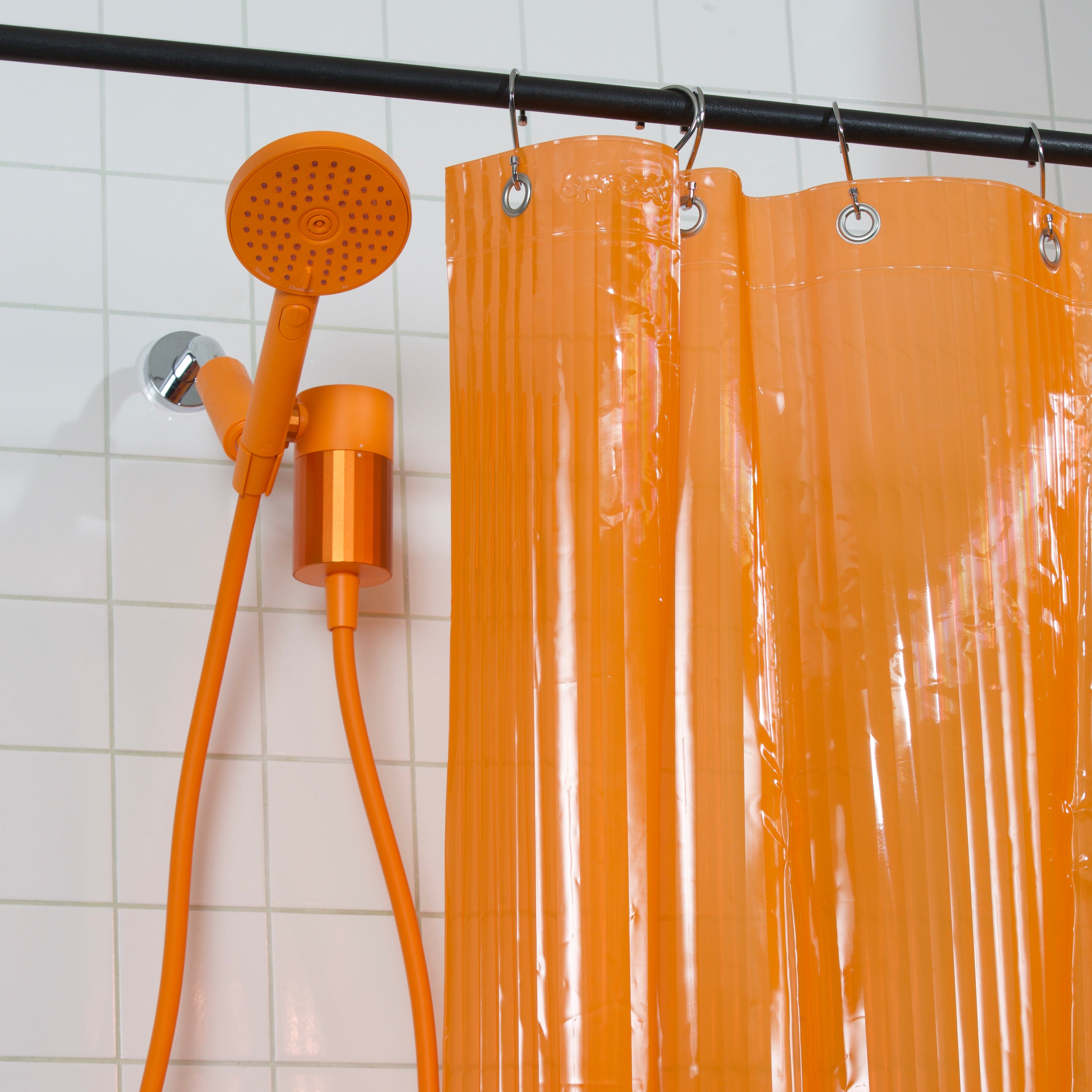 An orange shower curtain hanging from a black rod in a tiled bathroom, with an orange handheld shower head and hose mounted on the wall.